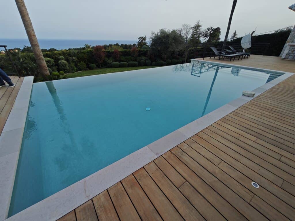 Piscine à débordement turquoise avec terrasse en bois, jardin luxuriant et vue panoramique sur la mer.
