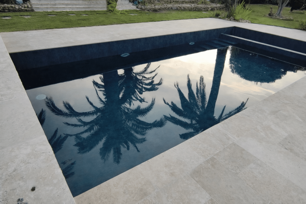 Piscine moderne au liner noir et margelles en travertin reflétant des palmiers sous un ciel voilé.