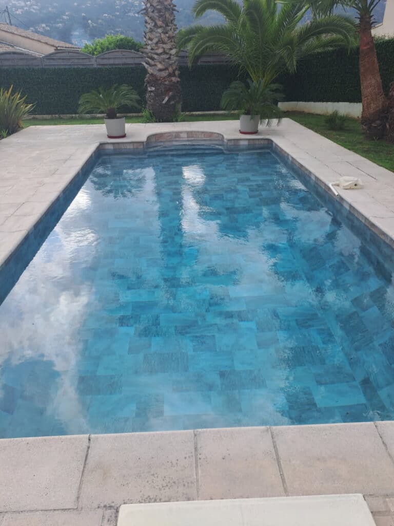Piscine rectangulaire en mosaïque bleue entourée de palmiers sur une terrasse en pierre claire.
