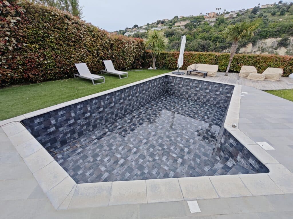 Piscine vide avec carrelage mosaïque gris et terrasse Piscine vide avec carrelage mosaïque gris, chaises longues sur gazon synthétique et vue sur une colline résidentielle.