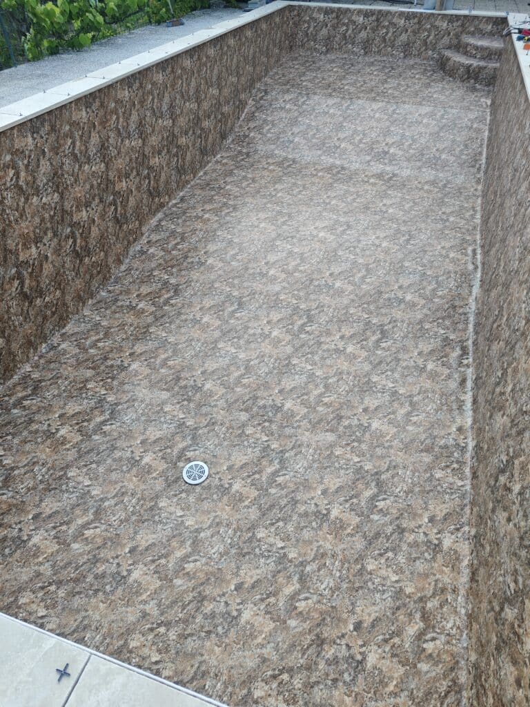 Liner piscine texturé effet pierre marron et beige, bassin vide avec margelles blanches et bonde de fond.
