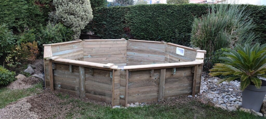 Piscine hexagonale en bois hors-sol installée dans un jardin paysager bordé de haies.