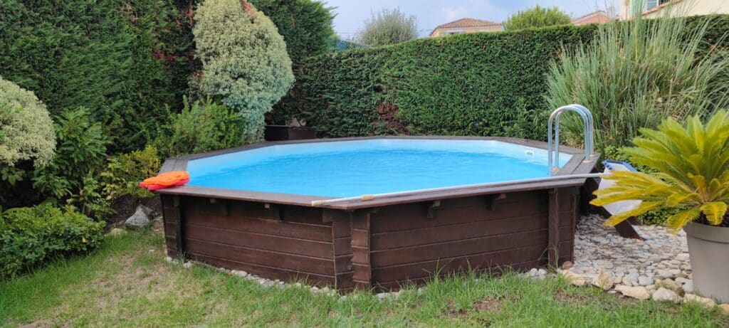 Piscine octogonale bois dans jardin avec haie et accessoires.
