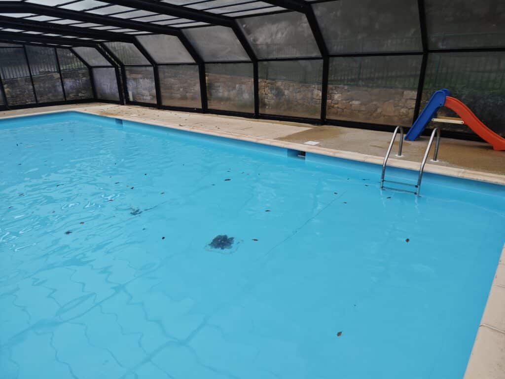 Piscine intérieure sous abri noir avec eau bleue, échelle inox et petit toboggan coloré.